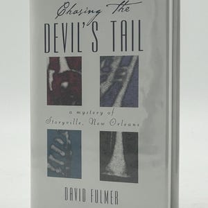 May include: A hardcover book titled "Chasing The Devil's Tail" by David Fulmer. The cover is light gray with the title in large black letters and four small, colorful images. The subtitle reads "a mystery of Storyville, New Orleans."