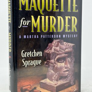 May include: A hardcover book titled "MAQUETTE for MURDER" by Gretchen Sprague. The cover features a stone bust and the text in yellow and white. The book is part of the "A Martha Patterson Mystery" series.