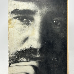 Peut inclure: Portrait en noir et blanc d'un homme avec une moustache et une barbe, imprimé sur une surface rectangulaire. L'image est un gros plan du visage de l'homme, avec un côté dans l'ombre. La surface a un aspect usé et vieilli.