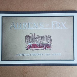 May include: A black framed poster with the text "Ahrens-Fox" in silver lettering. The poster features a red fire engine driving through a city skyline with the text "America's Foremost Fire Engine" below the image.