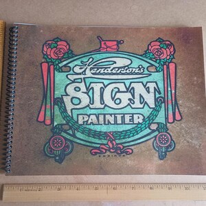 Op de afbeelding: Een spiraalgebonden notitieboek met een omslag met de tekst "Henderson's SIGN PAINTER" in een vintage ontwerp. Het ontwerp bevat rozen, decoratieve elementen en een turquoise en rood kleurenschema. Een houten liniaal ligt naast het notitieboek.