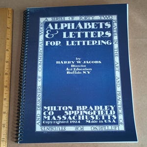 Puede incluir: Una cubierta azul de un libro titulado "Alphabets & Letters for Lettering" de Harry W. Jacobs. El libro es una serie de 42 láminas sobre el arte y la práctica de la rotulación para estudiantes y diseñadores de trabajos comerciales. Fue publicado por Milton Bradley Co. en Springfield, Massachusetts en 1924.