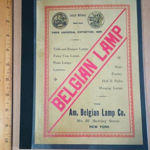 Op de afbeelding: Een vintage advertentie voor de Am. Belgian Lamp Co. in New York City. De advertentie heeft een rode diagonale banner met de tekst "BELGIAN LAMP" in witte letters. De advertentie heeft ook een gouden medaille met de tekst "GOLD MEDAL" en "Paris Universal Exposition, 1889".