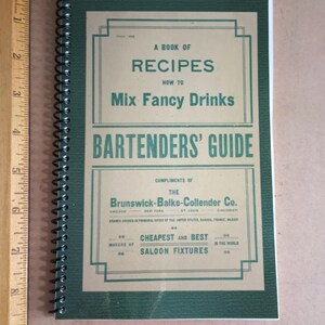 Könnte beinhalten: Ein grünes und braunes spiralgebundenes Buch mit dem Titel "A Book of Recipes How to Mix Fancy Drinks Bartenders' Guide" mit dem Text "Compliments of The Brunswick-Balke-Collender Co. Chicago New York St. Louis Cincinnati Branch Houses in Principal Cities of the United States, Canada, France, England Makers of Cheapest and Best Saloon Fixtures in the World."
