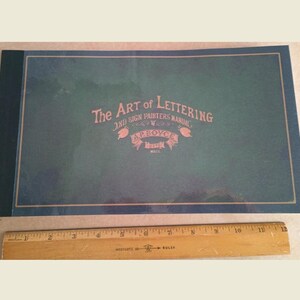 Op de afbeelding: De omslag van een groen boek getiteld "The Art of Lettering and Sign Painters Manual" door A.P. Boyce. Het boek is gebonden in donkerblauw met een gouden rand.