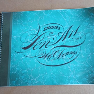 Op de afbeelding: Een spiraalgebonden boek getiteld "Studies in Pen Art" door W. Dennes. De omslag heeft een turquoise marmerontwerp met elegante scriptletters. Een houten liniaal ligt naast het boek.