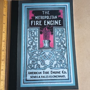 May include: A vintage book cover with a blue and silver design. The title is "The Metropolitan Fire Engine" with the text "American Fire Engine Co. Seneca Falls and Cincinnati." below the title.