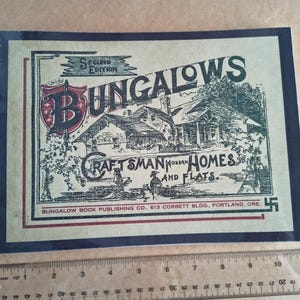 Puede incluir: Un libro encuadernado en espiral titulado "Bungalows, Craftsman Modern Homes and Flats". La portada presenta una ilustración de una casa y texto en un estilo vintage. El libro está sobre una superficie marrón claro con una regla.