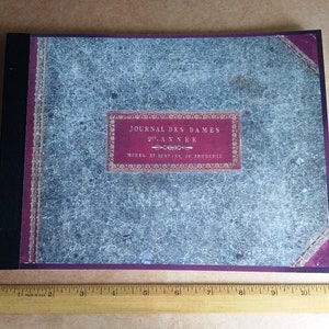 May include: A vintage, blue and red, bound journal with the title "Journal des Dames 20e Annee Modes et Dessins de Broderie" on the front cover.