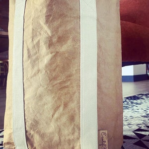 May include: A large, brown paper tote bag with two wide, cream-colored straps running vertically. The bag has a textured appearance and a small, rectangular tag with the text "Ecolif3". The bag is standing upright on a patterned rug.