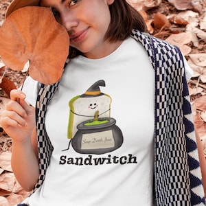 May include: White t-shirt with a cartoon sandwich wearing a witch hat and a cauldron that says "Soup Death Jinx". The word "Sandwitch" is printed below the graphic. The person is wearing a brown hat and gray pants.