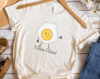 May include: A natural colored t-shirt with a graphic of a smiling egg with the text "Yolkie Dokie!" printed on it. The shirt is displayed on a wooden surface with a straw hat, sandals, and blue jeans.