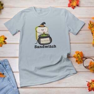 May include: Light blue t-shirt featuring a whimsical Halloween-themed graphic. The design includes a sandwich wearing a witch hat and holding a cauldron with the text "Sandwitch" below. The shirt is surrounded by autumn leaves.