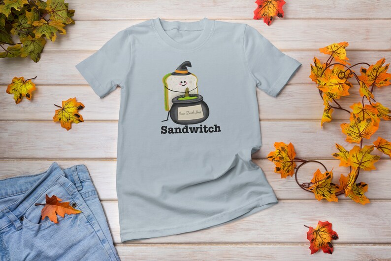 May include: Light blue t-shirt featuring a whimsical Halloween-themed graphic. The design includes a sandwich wearing a witch hat and holding a cauldron with the text "Sandwitch" below. The shirt is surrounded by autumn leaves.
