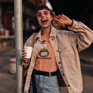May include: A person wearing a light orange t-shirt with a Halloween-themed graphic that says "Sandwitch". The person is also wearing a light pink jacket, blue jeans, and pink sunglasses, holding a white coffee cup.