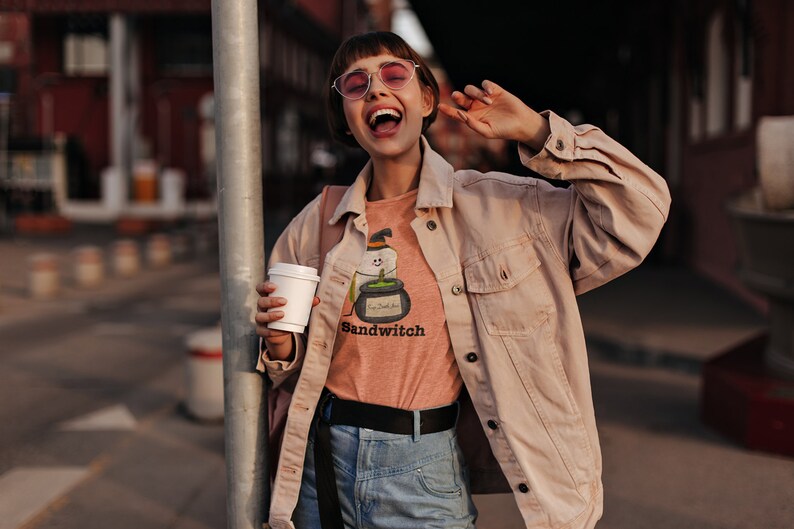 May include: A person wearing a light orange t-shirt with a Halloween-themed graphic that says "Sandwitch". The person is also wearing a light pink jacket, blue jeans, and pink sunglasses, holding a white coffee cup.