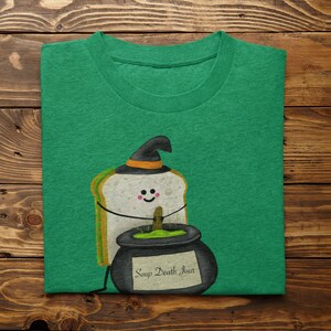 May include: Green t-shirt featuring a cartoon sandwich wearing a witch hat, with a smiling face and arms. A black cauldron with the text "Soup Death Jour" is next to the sandwich. The shirt is folded on a wooden surface.