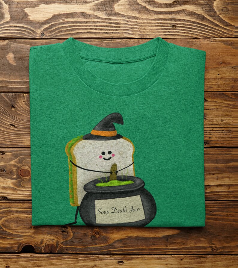 May include: Green t-shirt featuring a cartoon sandwich wearing a witch hat, with a smiling face and arms. A black cauldron with the text "Soup Death Jour" is next to the sandwich. The shirt is folded on a wooden surface.