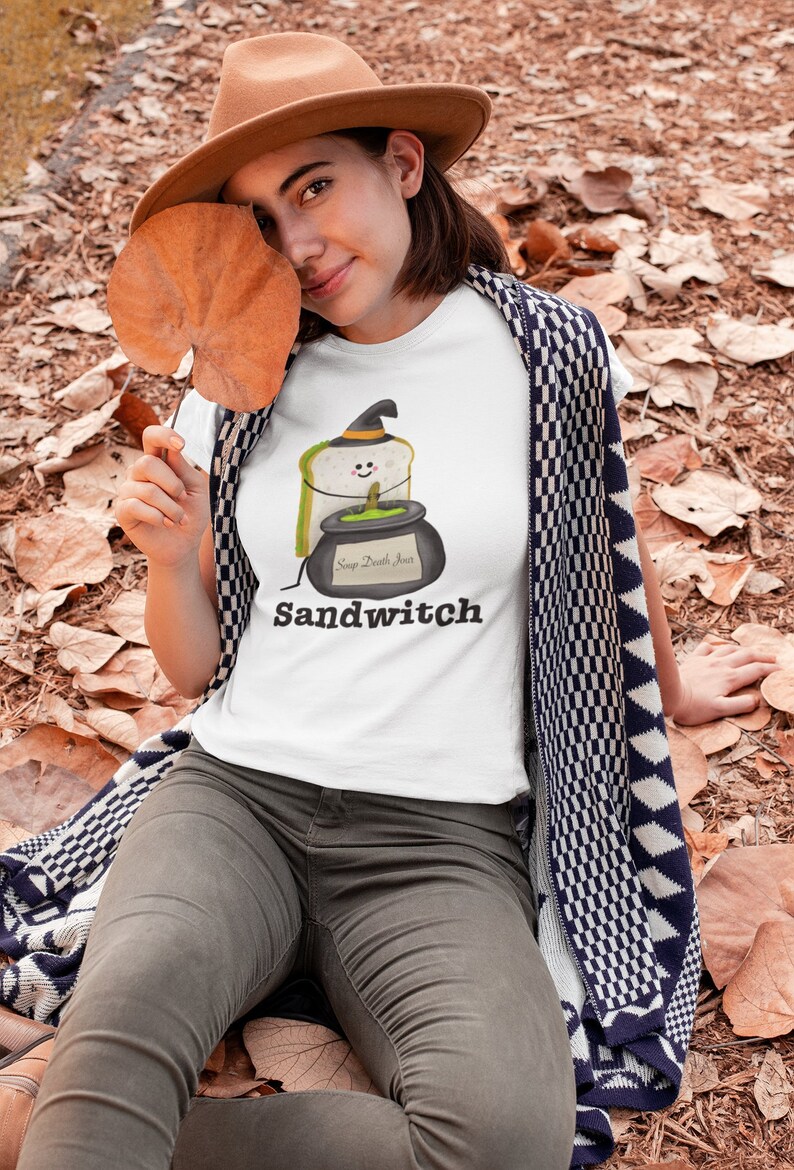 May include: White t-shirt with a cartoon sandwich wearing a witch hat and a cauldron that says "Soup Death Jinx". The word "Sandwitch" is printed below the graphic. The person is wearing a brown hat and gray pants.