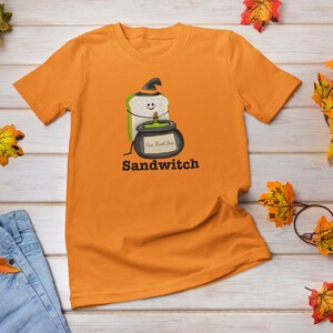 May include: Orange t-shirt featuring a cartoon sandwich wearing a witch hat and holding a cauldron with the text "Soup Death Juice". The word "Sandwitch" is printed below the graphic. The shirt is displayed on a wooden surface with autumn leaves.