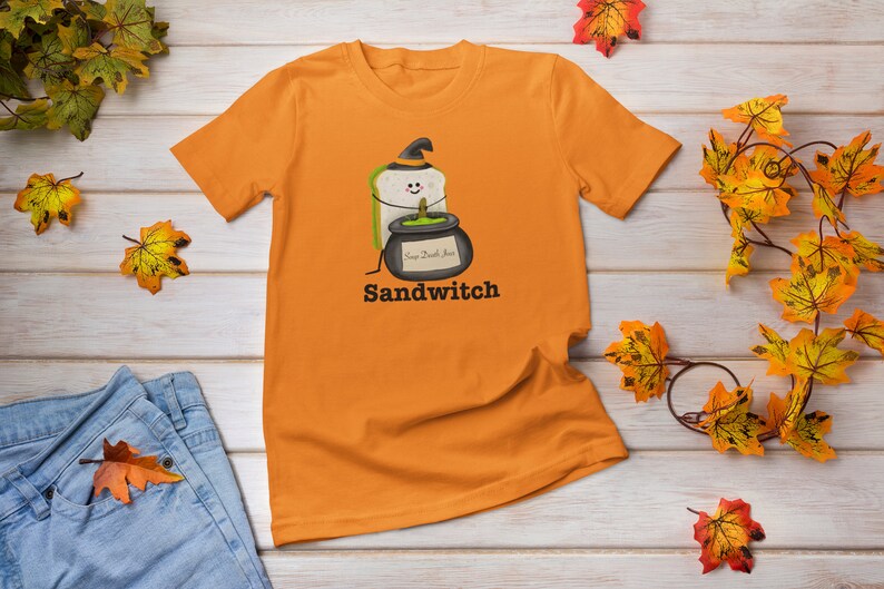 May include: Orange t-shirt featuring a cartoon sandwich wearing a witch hat and holding a cauldron with the text "Soup Death Juice". The word "Sandwitch" is printed below the graphic. The shirt is displayed on a wooden surface with autumn leaves.