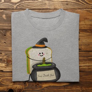 May include: A gray t-shirt featuring a cartoon sandwich wearing a witch hat and holding a cauldron. The cauldron has green liquid and the text "Soup Death Jour". The shirt is folded on a wooden surface.
