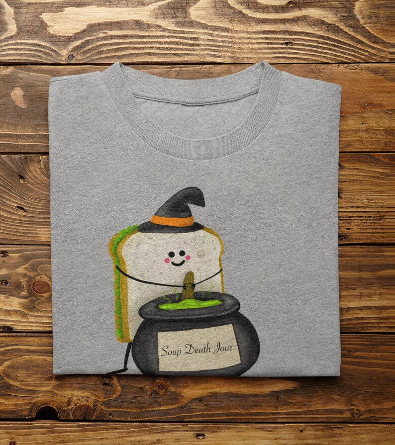 May include: A gray t-shirt featuring a cartoon sandwich wearing a witch hat and holding a cauldron. The cauldron has green liquid and the text "Soup Death Jour". The shirt is folded on a wooden surface.