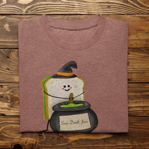 May include: A mauve t-shirt featuring a cartoon sandwich wearing a witch hat. The sandwich is next to a black cauldron with the text "Soup Death Jour". The design has a Halloween theme.