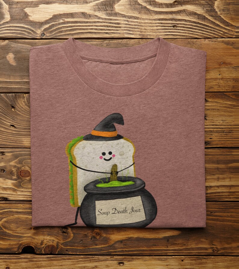 May include: A mauve t-shirt featuring a cartoon sandwich wearing a witch hat. The sandwich is next to a black cauldron with the text "Soup Death Jour". The design has a Halloween theme.