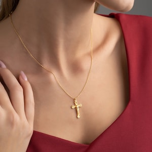 May include: A gold chain necklace with a delicate, ornate cross pendant. The necklace is worn by a person with a red shirt.