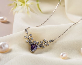 May include: A silver necklace with a purple gemstone deer pendant and clear crystal accents. The necklace is displayed on a white fabric background with white pearls.