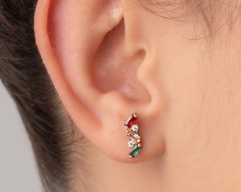 May include: A close-up of a person's ear with a small gold earring featuring red, green, and white gemstones.