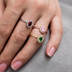 May include: Three silver rings with pear-shaped gemstones. The rings feature a red, pink, and green gemstone surrounded by a halo of smaller clear stones.