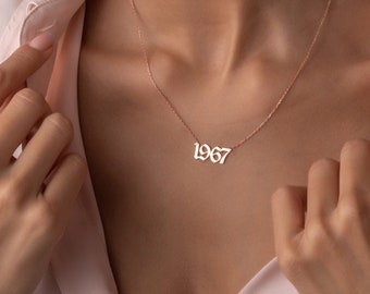 May include: A woman wearing a light pink shirt with a gold necklace that has the year 1967 on it.