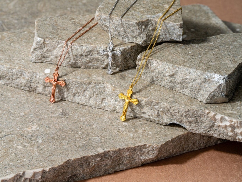 May include: Three gold, silver and rose gold necklaces with ornate cross pendants on a grey stone background.