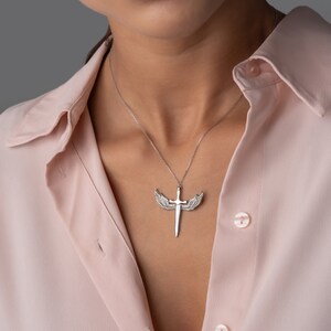 May include: A silver necklace with a pendant featuring a sword with wings. The pendant is on a silver chain and is worn by a person with a pink shirt.