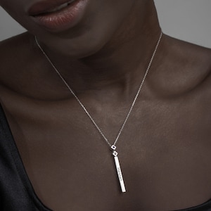 May include: A silver necklace with a long, thin pendant that says "Hannah" on it. The pendant is attached to a delicate chain with two small, square-shaped charms.
