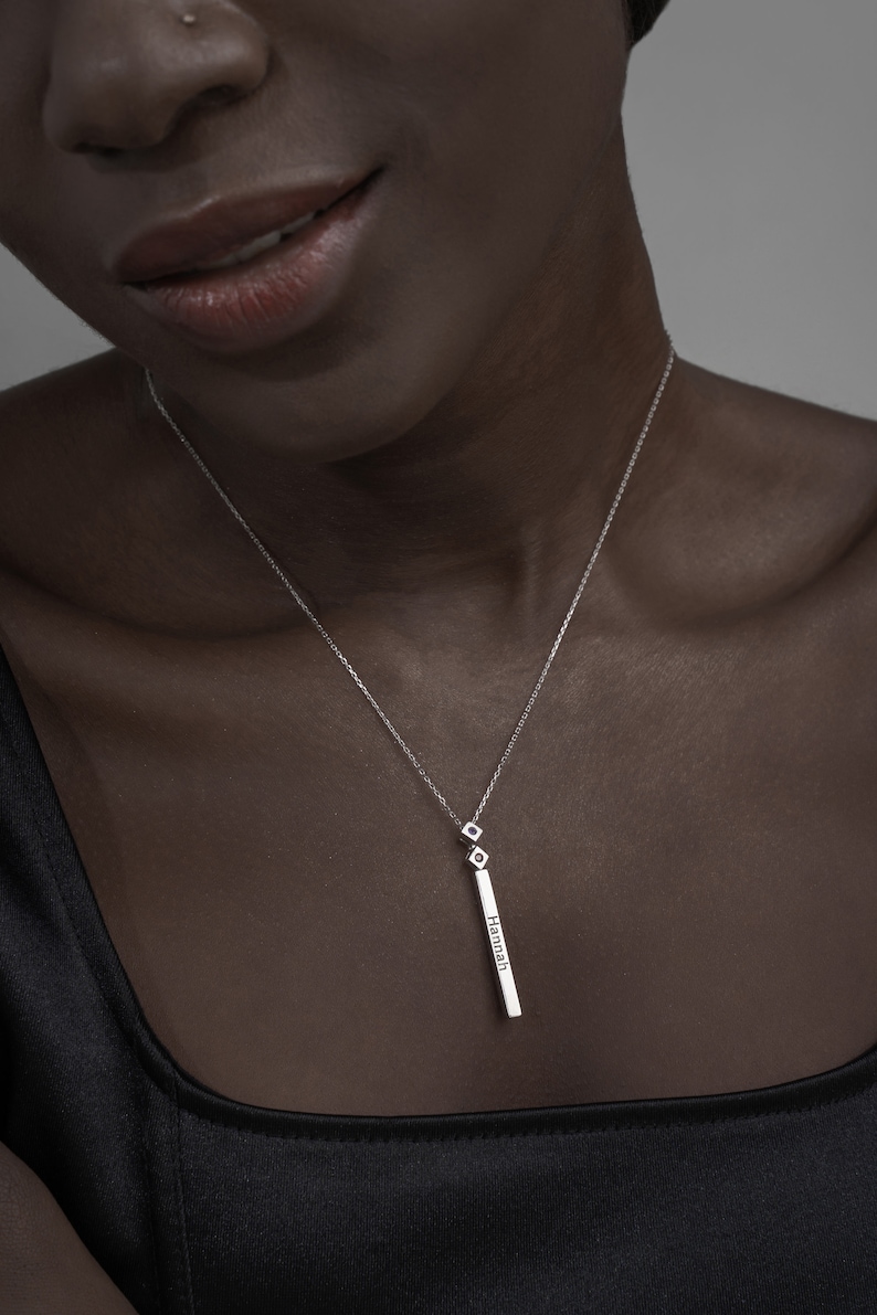 May include: A silver necklace with a long, thin pendant that says "Hannah" on it. The pendant is attached to a delicate chain with two small, square-shaped charms.
