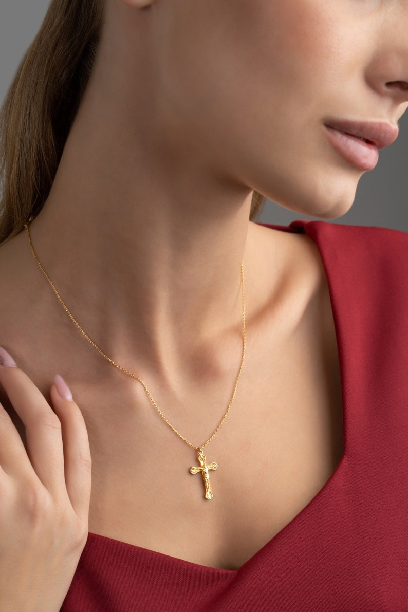 May include: A gold chain necklace with a delicate, ornate cross pendant. The necklace is worn by a person with a red shirt.