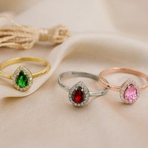 May include: Three rings with pear-shaped gemstones set in a halo of smaller stones. The rings are gold, silver, and rose gold. The gemstones are green, red, and pink.
