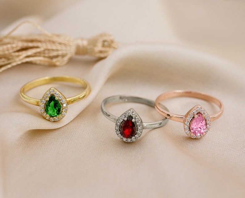 May include: Three rings with pear-shaped gemstones set in a halo of smaller stones. The rings are gold, silver, and rose gold. The gemstones are green, red, and pink.