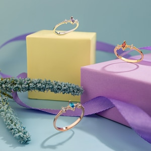 Puede incluir: Tres anillos de oro con piedras preciosas de diferentes colores sobre un fondo morado y amarillo. Los anillos son delicados y presentan un diseño en forma de corona.