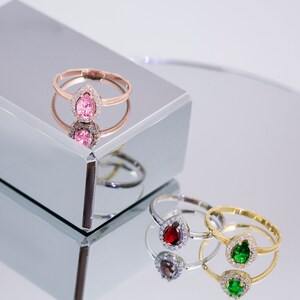 May include: Three rings with pear-shaped gemstones set in a halo of smaller stones. The rings are on a mirrored surface. The gemstones are pink, red, and green.