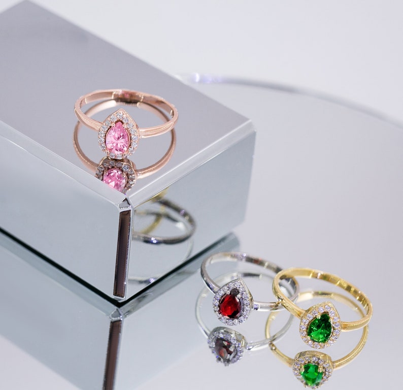 May include: Three rings with pear-shaped gemstones set in a halo of smaller stones. The rings are on a mirrored surface. The gemstones are pink, red, and green.
