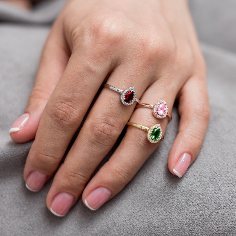 May include: Three silver rings with pear-shaped gemstones. The rings feature a red, pink, and green gemstone surrounded by a halo of smaller clear stones.
