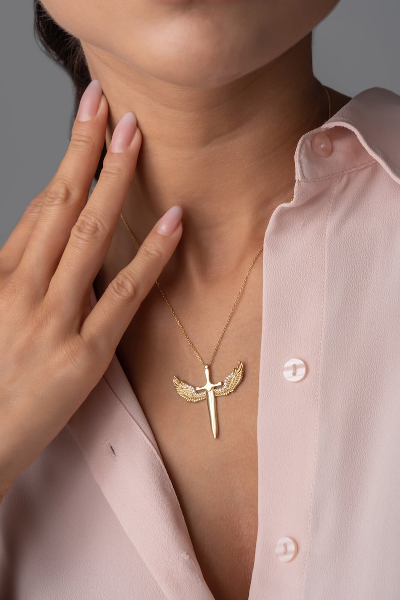 May include: A gold necklace with a pendant featuring a sword with wings. The wings are adorned with small, clear stones.
