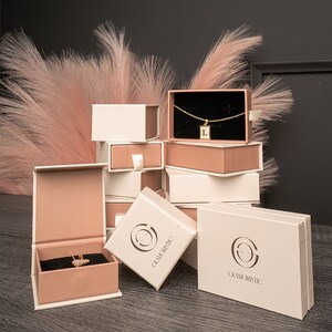 May include: Stack of small, white and pink gift boxes with the logo "Glamoristic" printed on the front. The boxes are arranged in a pyramid shape. The top box contains a gold necklace with a small pendant.