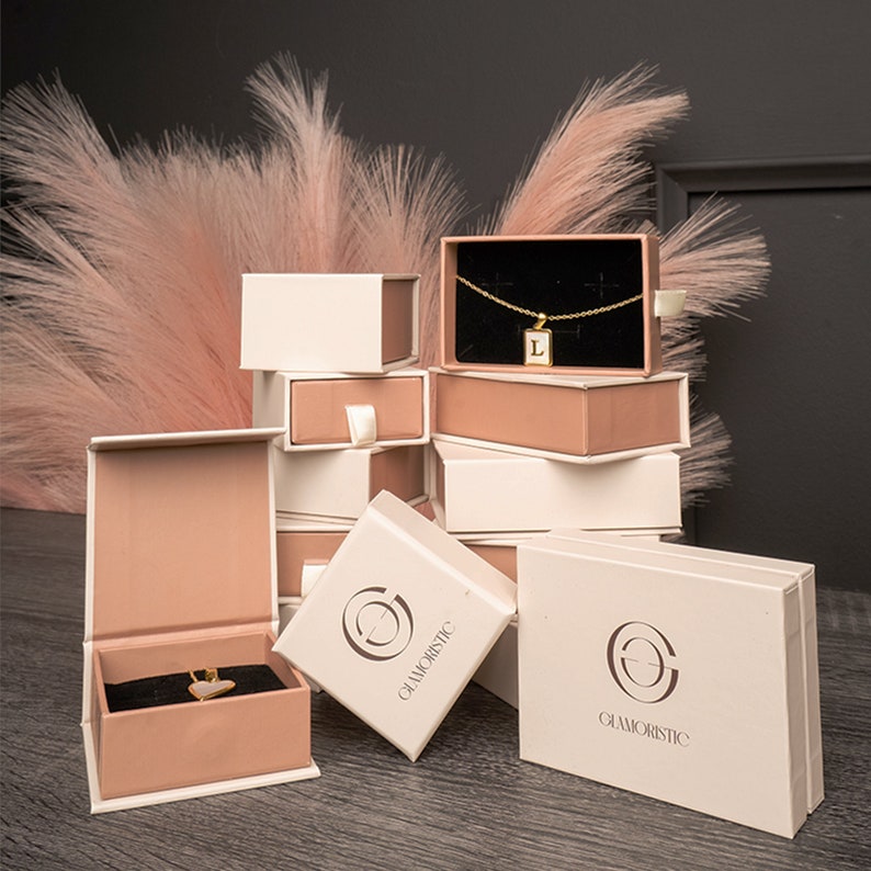 May include: Stack of small, white and pink gift boxes with the logo "Glamoristic" printed on the front. The boxes are arranged in a pyramid shape. The top box contains a gold necklace with a small pendant.