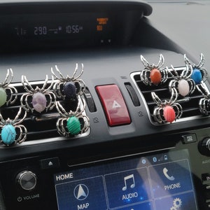 May include: Eight silver spider car air freshener clips with different colored gemstone accents. The spiders are hanging from the air vents of a car dashboard.