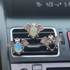 May include: Three silver bat car air fresheners with different colored glass stones. The bats are hanging from the air vent in a car. The stones are green, blue, and black.